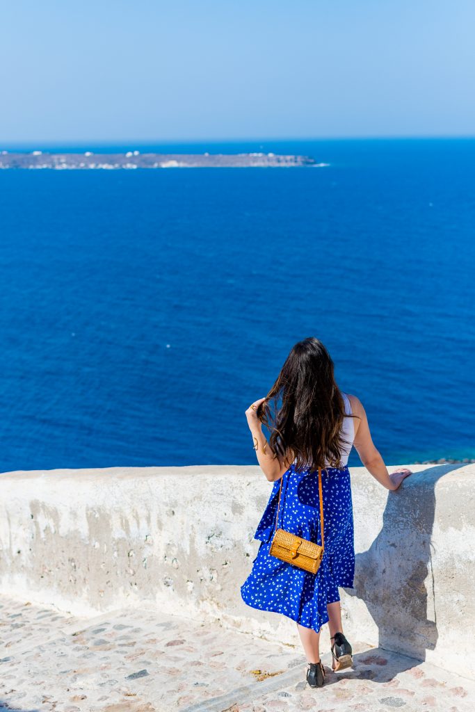 girl with long brown hair wearing a long royal blue skirt and white tank top overlooking the aegean sea from the stairway leading down to ammoudi bay in oia santorini greece