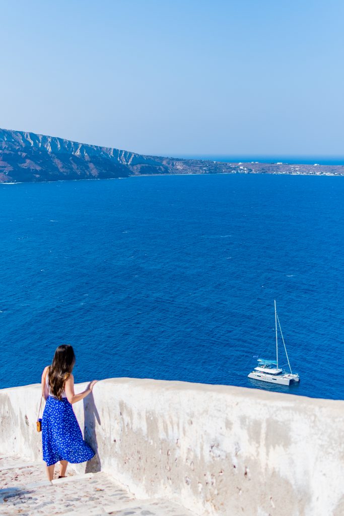 girl in blue Faithfull The Brand skirt and white tank top with long brown hair overlooking the agean sea from the stairway down to ammoudi bay in oia santorini greece