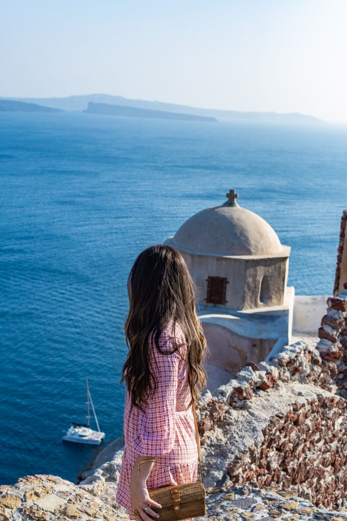 girl with long brown hair and red long sleeve dress standing on top of oia castle in santorini greece overlooking the big gray dome of the castle and the aegean sea