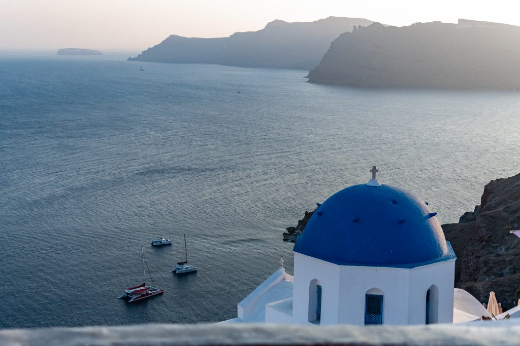 view of blue dome and aegean sea with sailboats from melenio cafe in Oia, santorini Greece