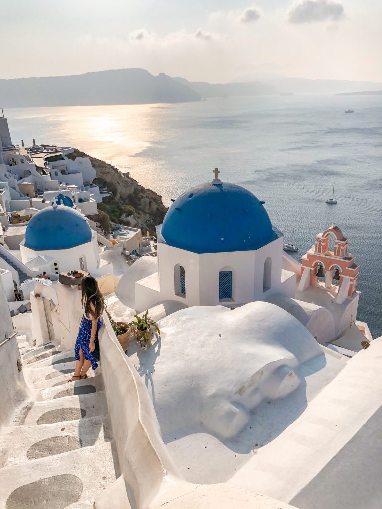 girl standing on stairs next to blue domes in oia, santorini greece with pink bell structure and ocean in the background