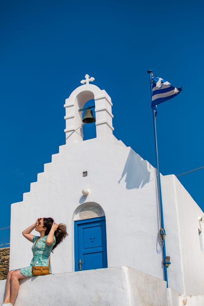 girl sitting on old white church with blue door and bell with greek flag flying, churches in mykonos, mykonos churches, best things to do in mykonos, things to see in mykonos