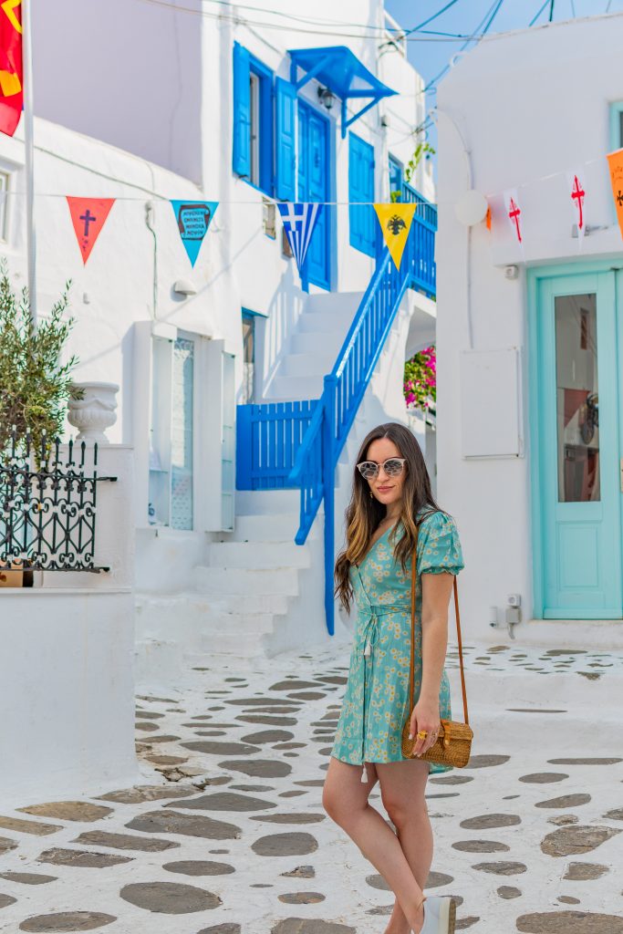 Girl posing in square in front of Church of Panachrantos (Panachra), panachra church in mykonos, greece, girl posing in front of church of panachra in mykonos with colorful flags hanging overhead with blue doors and stairways