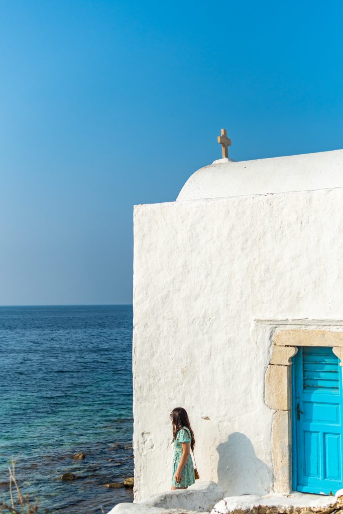 girl standing next to old church with a blue door next to the aegean sea in mykonos greece, best churches in mykonos