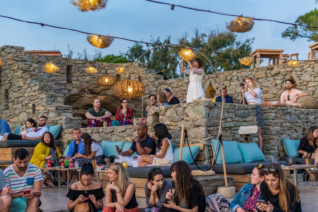 People hanging out and lounging at 180° sunset bar in mykonos greece, cocktail lounge in mikonos greece, sunset view from 180° Sunset Bar in Mykonos Greece overlooking Mykonos Town and the windmills. Where to watch the sunset in mykonos, the best things to do in mykonos greece