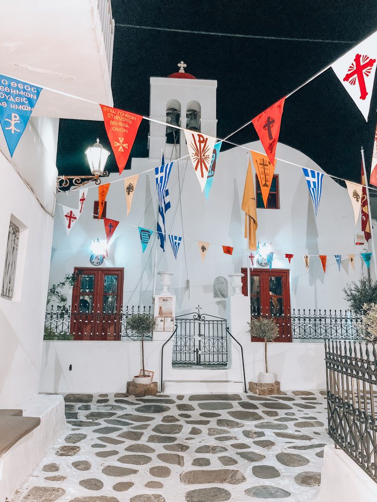 Church of Panachrantos (Panachra), panachra church in mykonos, greece, girl posing in front of church of panachra in mykonos with colorful flags hanging overhead and bright orange church door