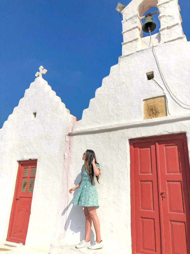 girl standing on old white church with red doors and bell, churches in mykonos, mykonos churches, best things to do in mykonos, things to see in mykonos