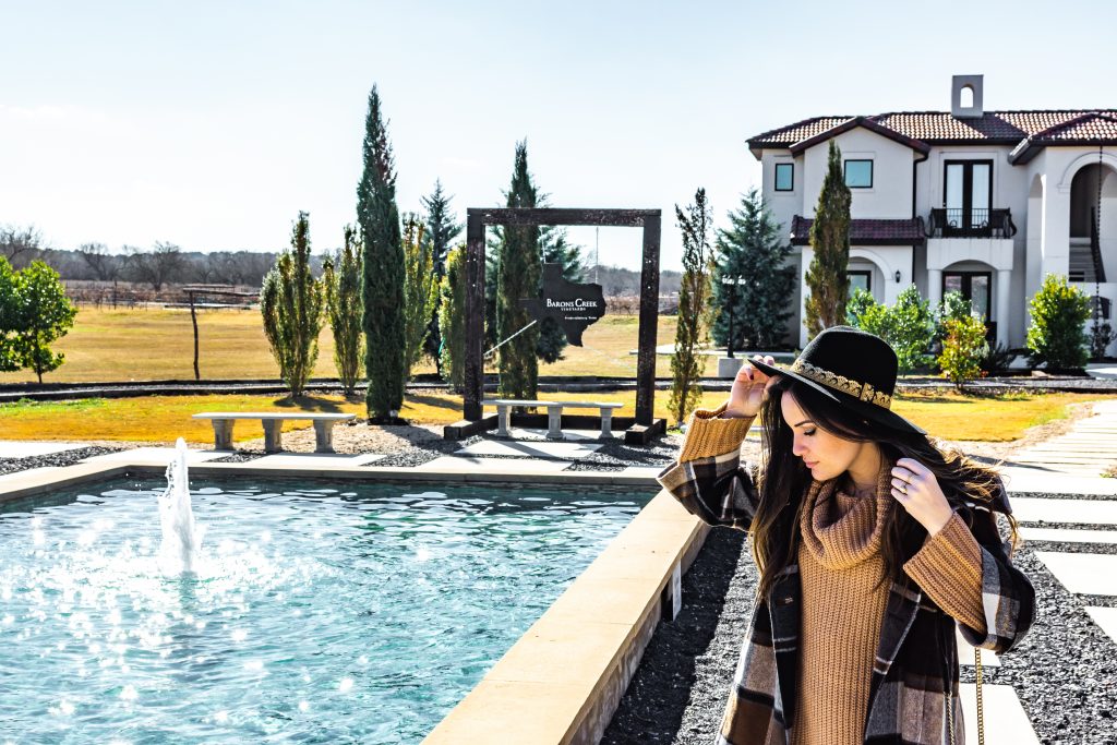 barons creek vineyards fredericksburg texas, winery in fredericksburg tx, tuscan style winery in fredericksburg with pool fountain and cottages behind winery, girl with sweater dress, coat and black hate at baron's creek vineyard